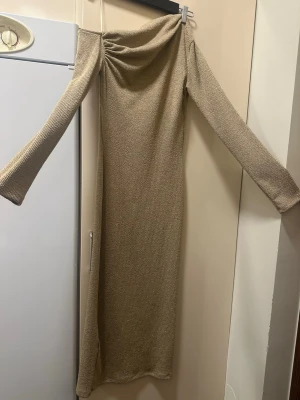 Guldig offshoulder långklänning från Zara - Snygg guldig långklänning med offshoulder-design och långa ärmar. Klänningen har en glittrig yta och ett elegant fall. Perfekt för dig som vill sticka ut med en unik look. Materialet känns mjukt och följsamt mot kroppen.