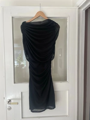 Svart omlottklänning med drapering - Elegant svart klänning med omlott och draperad design. Klänningen har en figurnära passform och är tillverkad i ett tunt, mjukt material som ger ett snyggt fall. Perfekt för dig som vill ha en stilren och trendig look.