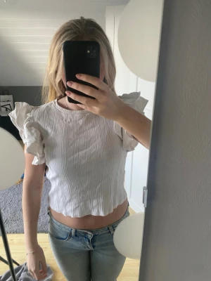 Vit volangblus med korta ärmar - Säljer en söt vit blus med volangdetaljer på axlarna och korta ärmar. Toppen har en lätt ribbad struktur och är croppad, vilket ger en trendig look. Perfekt att matcha med jeans eller kjol. Storlek 164
