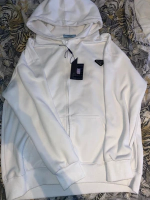 Vit hoodie med dragkedja från Prada - Köpt för 1300kr Snygg vit hoodie från Prada med dragkedja framtill och klassisk huva. Hoodien har en diskret ficka på magen och Pradas ikoniska trekantslogga på bröstet. Tillverkad i mjukt material som känns lyxigt och bekvämt. Perfekt för en clean och stilren look.