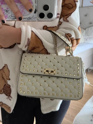 Vit Valentino Garavani handväska med gulddetaljer - Säljer en snygg vit rockstudd bag från Valentino Garavani med quiltat mönster och guldfärgade nitar. Väskan har ett  handtag och en kedjerem i guld som går att justera så att den blir längre, samt ett vridlås framtill. Kan bäras på olika sätt! Kan inte garantera äkthet (köpt på plick) men pris kan diskuteras🩷