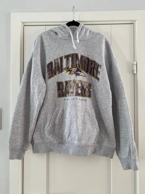 Grå Baltimore Ravens hoodie - Säljer en grå hoodie med stor Baltimore Ravens-logga och text i lila och gult på bröstet. Klassisk känguruficka framtill och vit dragsko i huvan. Perfekt för dig som gillar amerikansk fotboll och vill ha en sportig stil. Använd och har lite små noppor i övrigt i bra begagnat skick.  Mått Över bröstet: ca 60 cm Längd från axeln och ner: ca 65 cm  80% bomull och 20% polyester  Kommer från ett djur och rökfritt hem