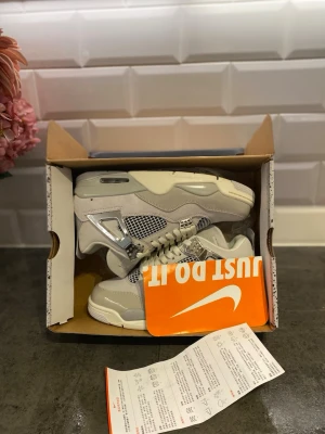 Nike Air Jordan 4 Retro Metallic Silver - Snygga Nike Air Jordan 4 Retro sneakers i vitt och ljusgrått med metallic silver detaljer. Skorna har meshpaneler, synlig Air-enhet i sulan och klassisk Jumpman-logga på hälen. Ovandelen är i mocka och läder, och de har en chunky sula för extra streetkänsla.