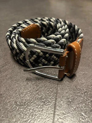 Svartvitt flätat bälte med läderdetaljer - Snyggt flätat bälte i svart och vitt med bruna läderdetaljer vid spännet och änden. Bältet har ett klassiskt metallspänne och passar perfekt till jeans eller chinos för en stilren look.