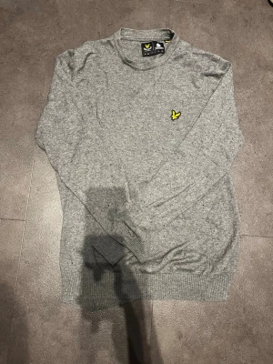Grå stickad tröja från Lyle & Scott - Snygg grå stickad tröja från Lyle & Scott med deras klassiska gula fågel-logga broderad på bröstet. Tröjan har rund halsringning, långa ärmar och ribbade muddar vid ärmslut och nederkant. Perfekt till lager på lager och enkel att matcha.