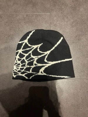 Spi5der mössa  - Säljer en svart mössa med ett vitt spindelnätsmönster som täcker ena sidan. Mössan är stickad och har en klassisk, något slouchy passform. Perfekt för dig som vill ha en edgy och unik accessoar till hösten eller vintern.