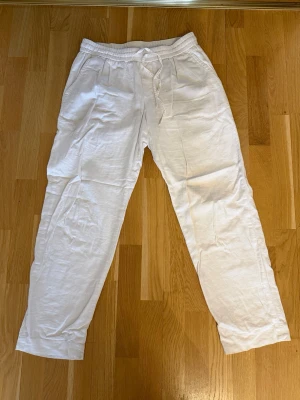 Vita linnebyxor med dragsko - Säljer ett par vita linnebyxor med avslappnad passform och dragsko i midjan. Byxorna har sidofickor fram och två fickor bak. Perfekta för en chill och stilren look. Materialet är lätt och luftigt linne.