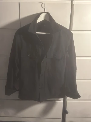 Svart overshirt från Uniqlo - Stilren svart overshirt från Uniqlo med klassisk krage och knäppning framtill. Två bröstfickor med knappar ger en cool och praktisk look. Perfekt att bära som lager över t-shirt eller hoodie. Materialet är slätt och känns bekvämt.