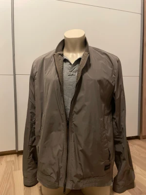 Dondup vindjacka - Stilren beige vindjacka med hel dragkedja framtill och ståkrage. Jackan har långa ärmar, elastisk nederkant och diskret logotypmärke vid fickan. Tillverkad i lätt och vindtätt material, perfekt för blåsiga dagar.