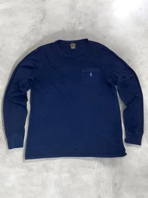 Marinblå långärmad t-shirt Polo Ralph Lauren - Snygg marinblå långärmad t-shirt från Polo Ralph Lauren med klassisk rund hals och bröstficka. Ikoniska broderade loggan i ljusblått på fickan. Tillverkad i mjuk bomull för skön känsla och stilren look.