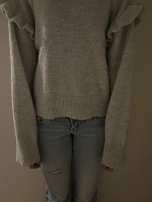 Grå stickad tröja med volang - Mysig grå stickad tröja med volangdetaljer vid axlarna och långa ärmar. Tröjan har en rak passform och ribbstickade muddar vid ärmslut och nederkant. 