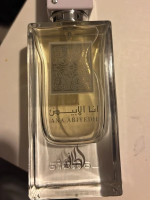 Ana Abiyedh parfym från Lattafa - Ana Abiyedh parfym från Lattafa i en elegant, genomskinlig glasflaska med silverdetaljer och orientaliskt mönster på etiketten. Flaskan har en stilren, rektangulär form och vitt lock. Doften är känd för sina fräscha och träiga noter.