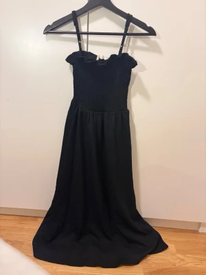 Svart långklänning med smock - Svart långklänning med smala axelband och smockad överdel. Klänningen har volangdetalj vid bysten och ett mjukt, flytande tyg som ger en elegant siluett. Perfekt för dig som gillar stilrena och klassiska plagg.