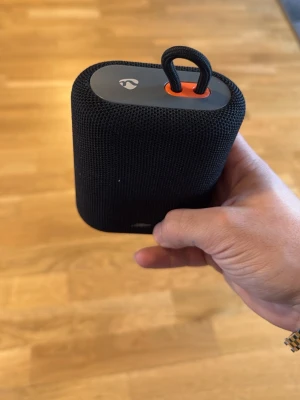  högtalare - Smidig svart bärbar högtalare från Linocell med tygklädd yta och orange bärögla. Högtalaren har USB-C laddning och tydliga knappar för volym och ström. Perfekt för att ta med sig överallt tack vare sin kompakta form och robusta material.