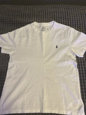 Vit t-shirt från Polo Ralph Lauren - Klassisk vit t-shirt från Polo Ralph Lauren storlek 10-12 år/ 155-165.med den ikoniska broderade loggan på bröstet. T-shirten har rund hals, korta ärmar och är gjord i mjuk bomull för en skön känsla. Perfekt basplagg till garderoben.