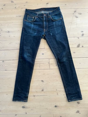 Mörkblå jeans från Nudie - Snygga mörkblå slimfit jeans från Nudie med klassiska fem fickor och kontrastsömmar. Jeansen har en slim passform och är tillverkade i slitstark denim med en cool tvätt. Perfekta för dig som gillar en stilren och modern look. Jeansen är gjorda i ett dry denim material vilket gör att de får en speciell Fade vid användning. Jeansen är i storlek 31W 32L men sitter mer som en 30W 32L. Modell är lean Dean. Vid frågor eller funderingar är det bara att säga till Mvh Olof.