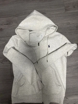 Ljusgrå hoodie från Polo Ralph Lauren - Snygg ljusgrå hoodie med dragkedja från Polo Ralph Lauren. Klassisk design med huva och dragsnören, två fickor framtill och den ikoniska mörkblå loggan broderad på bröstet. Tillverkad i mjukt bomullsmaterial för maximal komfort.
