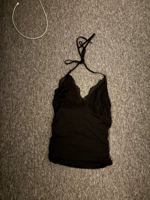 Svart spetstopp med halsterneck - Snygg svart topp med halterneck och djup urringning. Toppen är använd vid enstaka tillfällen och är i storlek S 