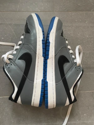 Nike Dunk Low grå/svarta sneakers - Nike Dunk Low sneakers i grått och svart läder med vit sula och blå detaljer undertill. Klassisk svart swoosh på sidan och vita skosnören. Skön och stilren modell som passar till det mesta. Perfekt för dig som gillar streetwear och sneakers. Nypris ca 1600 kr