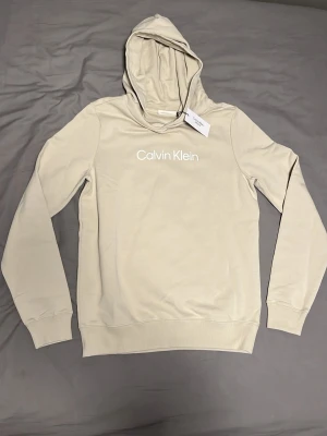 Beige hoodie från Calvin Klein Jeans - Snygg beige hoodie från Calvin Klein Jeans med vit logga tryckt på bröstet. Klassisk modell med huva och ribbade muddar vid ärmar och nederkant. Tillverkad i mjukt bomullsmaterial som känns skönt mot huden. Perfekt för en avslappnad och trendig look.