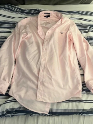 Ljusrosa skjorta från Gant - Säljer en ljusrosa skjorta från Gant, modell The Archive Oxford. Skjortan har klassisk krage, knappar framtill och en liten broderad logga på bröstet. Tillverkad i mjuk bomull och har lång ärm. Perfekt för en clean och stilren look.