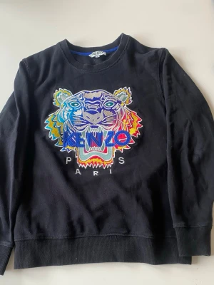 Svart sweatshirt med tiger från Kenzo - Cool svart sweatshirt från Kenzo med ikonisk broderad tiger i flera färger och texten 'KENZO PARIS' på bröstet. Tröjan har rund halsringning och ribbade muddar. Perfekt statement-plagg för dig som gillar streetwear och färgstarka detaljer.
