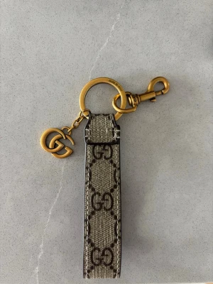nyckelring med GG-logga - Säljer en snygg nyckelring från Gu*** med klassiskt GG-mönster i beige och brunt. Metall i guldfärg och GG-hänge. Insidan har den ikoniska gröna och röda randen. Perfekt accessoar för dig som gillar lyxiga detaljer. Väldigt sparsamt använd, användes max i 1 vecka 