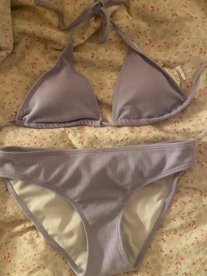 Lila bikini - Supersnygg ljuslila bikini från NA-KD med ribbad struktur. Överdelen har trekantsform och knyt i nacken, medan underdelen är klassisk med låg midja. Perfekt för lata dagar på stranden eller poolen.