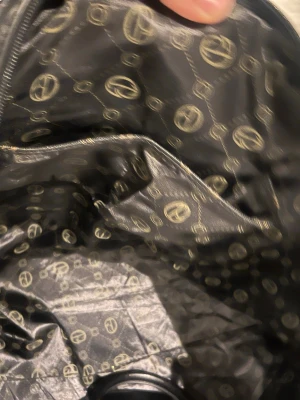 Svart resväska från Giulia Pieralli  - Snygg svart resväska från Giulia Pieralli med ikoniskt mönstrat innerfoder i beige och svart. Väskan har en praktisk innerficka med dragkedja och är tillverkad i slitstarkt syntetmaterial. Perfekt för dig som vill resa med stil och ha ordning på dina grejer.