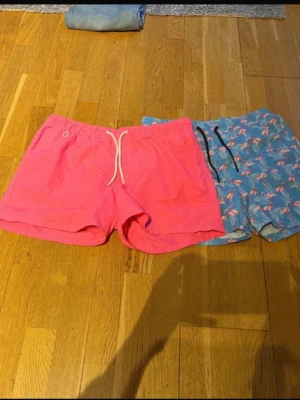 Två par badbyxor, rosa & flamingo - Säljer två par badbyxor, ett par i helrosa med vit dragsko och ett par ljusblå med flamingomönster och svart dragsko. Båda har korta ben och elastisk midja, perfekta för stranden eller poolen.fint skick! De rosa är från newyorker och flamini från Jack and jones 