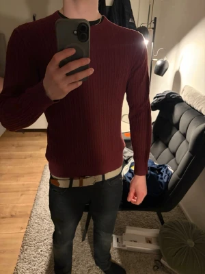 Vinröd kabelstickad tröja herr (Riley) - Säljer en vinröd kabelstickad tröja från Riley med rund hals och långa ärmar. Tröjan har ett klassiskt mönster och sitter snyggt på kroppen. Perfekt för dig som gillar stilrena och tidlösa plagg.