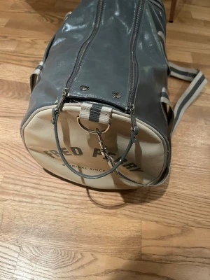 Grå och beige duffelväska Fred Perry - Snygg duffelväska från Fred Perry i grått och beige konstläder med rund form. Väskan har justerbar axelrem med randigt mönster och dragkedja upptill. Perfekt för gymmet eller som weekendbag. Märkeslogga tryckt på sidan.
