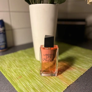 Liberty Life parfym Manhattan - Snygg parfymflaska från Liberty Life med doften Manhattan. Flaskan är rektangulär i genomskinligt glas med en svart, snedställd kork och innehållet har en varm orange ton. Helt ny