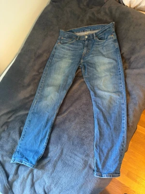 Blå Levi's jeans  - Klassiska blå Levi's jeans med raka ben. Jeansen har en snygg tvätt med slitningar på låren och knäna för en avslappnad look. Tillverkade i bomull med Levi's ikoniska läderpatch bak i midjan. Bältet har färgat av sig på lappen som syns i sista bilden. Utöver det har den inga defekter 