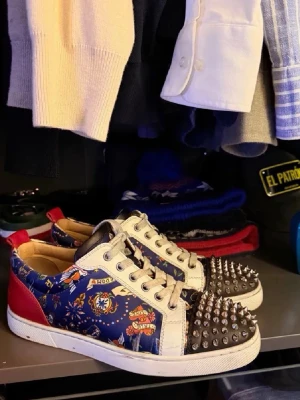 Christian Louboutin  sneakers - Äkta Storlek 41-42, Ok skick                                                          Unika sneakers från Christian Louboutin med blått mönstrat tyg, vita detaljer, röda hälpartier och signaturröda sulor. Tåpartiet är täckt av silverfärgade nitar för en edgy look. Skorna har vita snören och metalliska öljetter.
