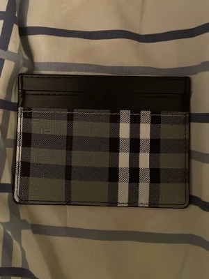 Rutig kortfodral från Burberry - Snyggt kortfodral från Burberry i svart och grått med klassiskt rutmönster och vita detaljer. Tillverkat i slätt läder med flera kortfack på båda sidor. Perfekt för dig som vill ha en stilren och lyxig accessoar i fickan.