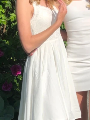 Vit spets studentklänning med v-ringning  - Söt vit klänning från Chiara Forthi med spetsdetaljer upptill och v-ringad hals. Kjolen är utsvängd och kort, vilket ger en luftig känsla. Klänningen är ärmlös och har dragkedja i ryggen. Perfekt för sommarens alla tillfällen.