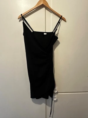 Svart omlottklänning  - Säljer en svart kort omlottklänning från Divided med smala axelband och lätt rynkad detalj på sidan. Klänningen har en figurnära passform och är tillverkad i mjukt syntetmaterial. Perfekt för dig som gillar minimalistisk och stilren design.