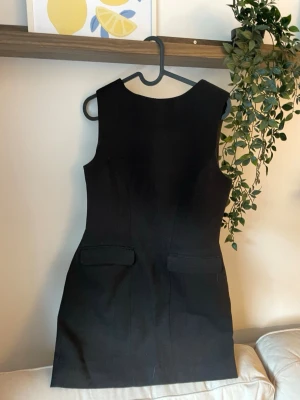 Svart ärmlös klänning med fickor från NA-KD - Stilren svart klänning i figurnära modell med ärmlös design och rund halsringning. Klänningen har två dekorativa fickor framtill och är tillverkad i ett slätt tyg som ger en klassisk look. Perfekt för dig som gillar minimalistisk stil.