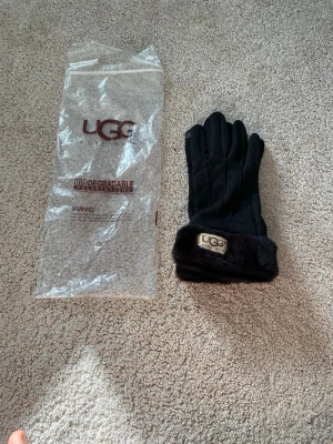 Svarta pälsfodrade handskar från UGG - Snygga svarta handskar från UGG med mjukt pälsfoder vid mudden och en beige logotyp på framsidan. Handskarna har en klassisk form och är perfekta för kalla dagar. Kommer med originalpåse.
