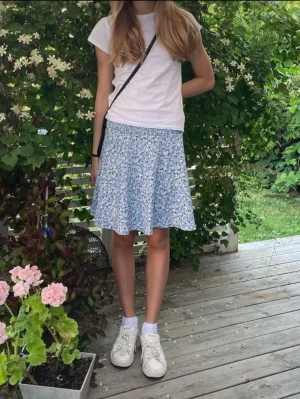 Blommig blå kortkjol i polyester  - Söt blå kortkjol med vitt och svart blommönster. Kjolen har en lätt utställd form och är gjord i mjukt polyester, perfekt för varma dagar. Enkel att matcha med en t-shirt eller linne för en avslappnad look.