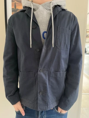 Mörkblå overshirt från Filippa K - Stilren mörkblå overshirt med tre utanpåliggande fickor och knäppning framtill. Overshirten har en klassisk krage och raka ärmar. Tillverkad i ett mjukt bomullstyg som ger en avslappnad look. Perfekt att bära över en hoodie eller t-shirt. Mitt pris 749kr