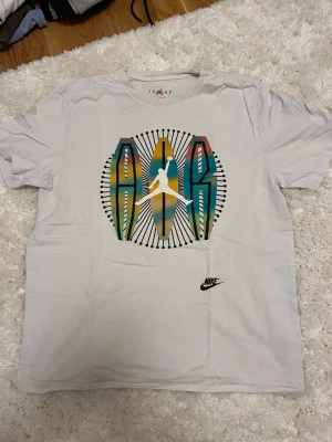 Vit Nike Air Jordan t-shirt - Säljer en vit t-shirt från Nike med stort färgglatt Air Jordan-tryck på bröstet och klassisk Jumpman-logga. T-shirten har rund hals och korta ärmar. Materialet känns mjukt och skönt, perfekt för en avslappnad stil.