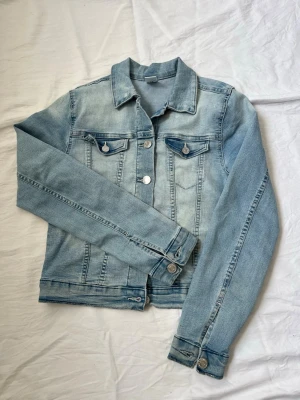 Ljusblå jeansjacka - Snygg ljusblå jeansjacka med silverfärgade knappar och två låssas bröstfickor. Jackan har en klassisk krage och raka sömmar för en tidlös look. Perfekt att slänga över en hoodie eller t-shirt för en avslappnad stil. Lite missfärgad på insidan av jackan. För flickor 11-12 år. Diskuterar gärna pris! ✨