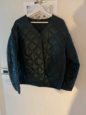 Svart quiltad vindjacka från Gina Tricot - Stilren svart vindjacka från Gina Tricot med quiltat mönster och V-ringad hals. Jackan har en lätt glansig yta och raka ärmar. Perfekt för lager-på-lager och enkel att matcha med olika outfits.
