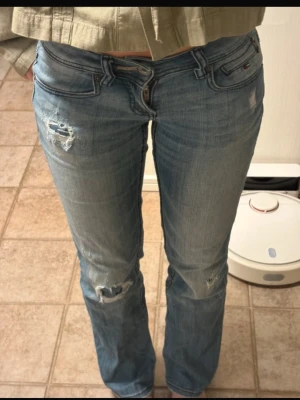 Ljusblå slitna bootcut jeans - Säljer ett par ljusblå jeans med slitningar och hål på låret och knät. Modellen är bootcut med låg midja och klassiska fem fickor. Jeansen har snygga detaljer och kontrastsömmar. Perfekta för en avslappnad och trendig look. Midjemått 37cm innerbenslängd 78cm