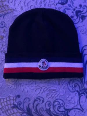 Svart Moncler mössa med randig kant - Snygg svart mössa från Moncler med uppvikt kant och klassiska ränder i blått, vitt och rött. Framtill sitter Monclers ikoniska logotyp. Perfekt accessoar för dig som vill ha en stilren och trendig look.