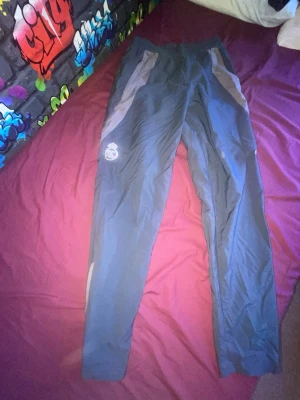 Adidas träningsbyxor grå - Säljer ett par grå träningsbyxor från Adidas med ljusgrå detaljer längs sidorna. Byxorna har elastisk midja och Adidas-logga på låret. Perfekta för träning eller chill. Materialet är lätt och andas bra.