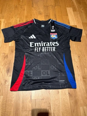 Lyon 24/25 away kit Cherki #18 - Snygg svart matchtröja från Olympique Lyonnais med Cherki 18 på ryggen. Tröjan har röda och blå detaljer på axlar och sidor, samt klubbmärke och Adidas-logga på bröstet. Tillverkad i lätt och ventilerande material, perfekt för fotboll eller träning.