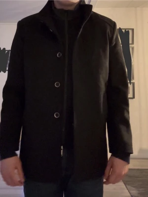 Svart klassisk rock med ståkrage - Stilren svart rock med rak passform och ståkrage. Rocken har diskreta knappar framtill och är tillverkad i ett slätt tyg som ger en elegant look. Perfekt för dig som gillar minimalistisk och tidlös stil.